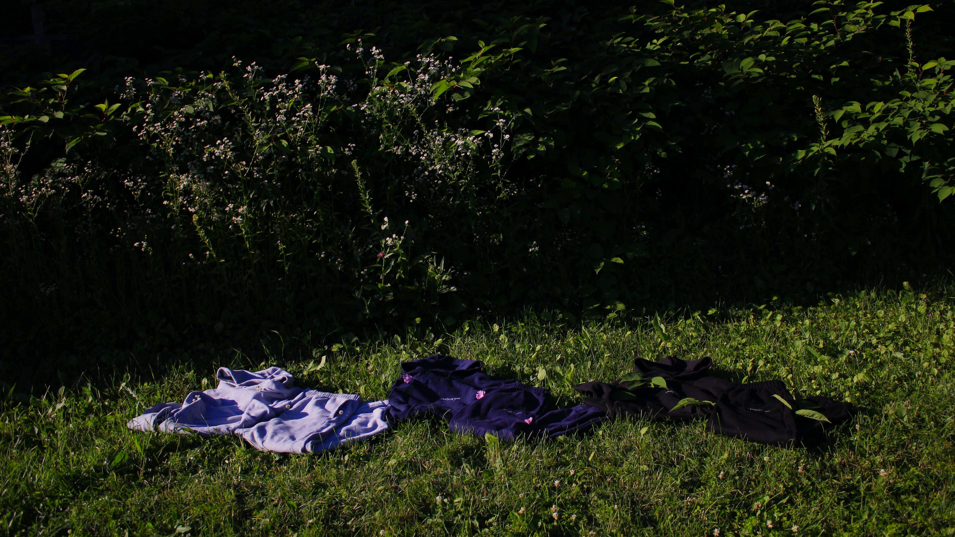 Three folded garments laid out on grass outdoors: a light grey set, a dark navy set, and a black set, with dense green bushes in the background.